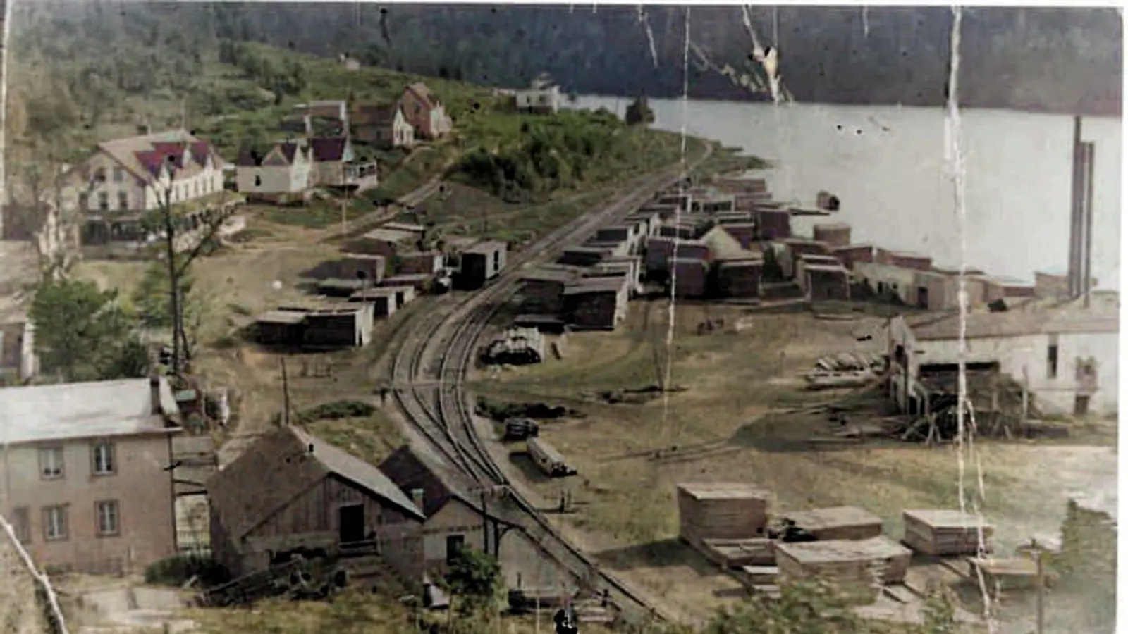 Photographie ancienne colorisée d'un village au bord d'un lac, avec une voie ferrée, des piles de planches et une scierie